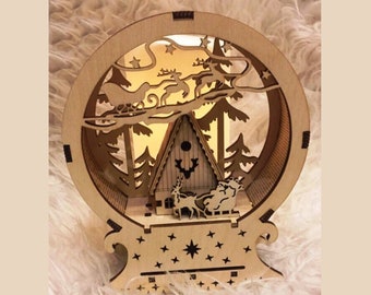 Può includere: Un ornamento natalizio in legno con un design a intaglio che rappresenta una scena invernale. L'ornamento presenta una piccola casa, alberi e renne. L'ornamento è illuminato dall'interno, creando un bagliore caldo.