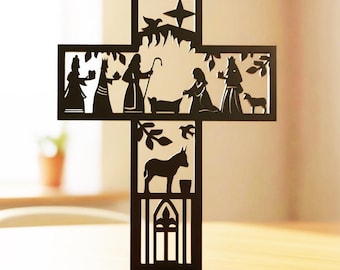 Peut inclure: Une croix noire plate découpée présentant une scène de nativité détaillée. La partie supérieure représente des personnages, une étoile et une crèche, tandis que la partie inférieure montre un âne, du feuillage et une arche en silhouette.