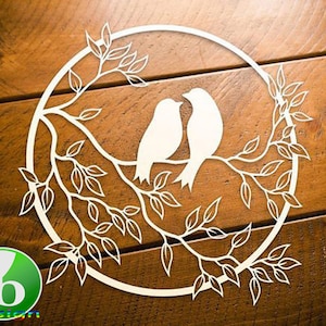 May include: A white paper cut-out depicting two birds on a branch, enclosed within a circular frame. The design features intricate leaves and branches, set against a wooden backdrop. The logo "ib design" is visible in the lower left corner.