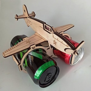 Könnte beinhalten: Holz Flugzeug-förmiger Getränkehalter mit zwei Getränkedosen. Das Flugzeug ist in Tarnfarben lackiert und hat einen roten Stern an der Seite. Die Dosen sind teilweise sichtbar und eine ist grün und schwarz, die andere ist rot.