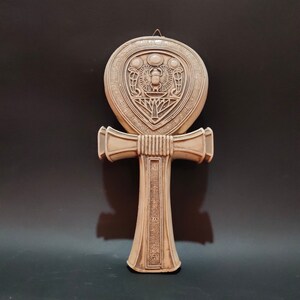 May include: A beige ceramic Ankh, an ancient Egyptian symbol of life, with intricate carvings and a scarab beetle design.