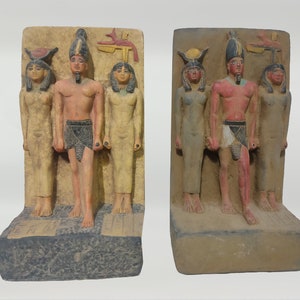 May include: Two Egyptian-style bookends, each featuring a carved relief of three figures. The figures are painted in muted tones of beige, red, and black. The bookends have a rectangular base and a textured background.