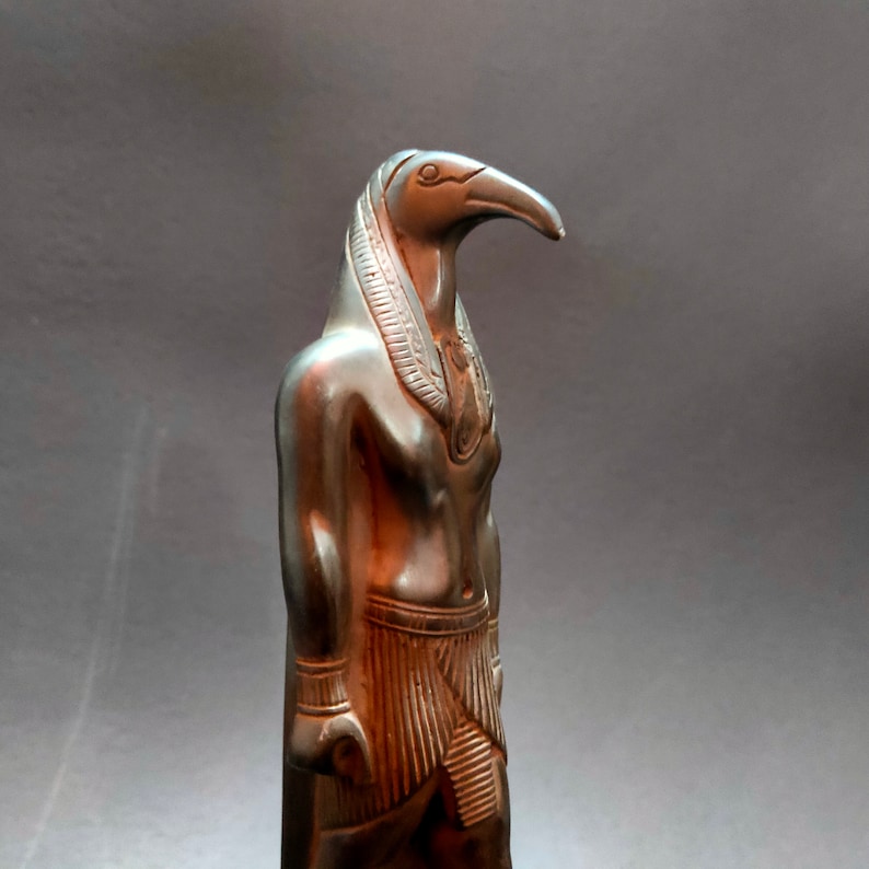 May include: A dark bronze statue of an Egyptian god with a bird head and human body. The statue is standing upright with its arms at its sides.