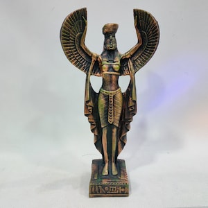 May include: A gold and green statue of an Egyptian goddess with outstretched wings. The goddess is wearing a headdress and a long, flowing robe. The statue is standing on a square base with hieroglyphics.