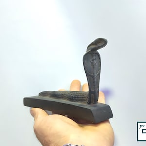 以下が含まれることがあります： 頭を持ち上げた、詳細なデザインが施された黒いコブラの像。像は黒い木製の台座の上に置かれています。