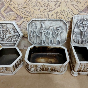 Könnte beinhalten: Vier dekorative Boxen mit ägyptischen Motiven. Die Boxen sind weiß mit erhabenen Details. Eine Box zeigt eine Kutschen-Szene, eine andere den Kopf eines Pharaos und zwei Boxen zeigen Szenen mit ägyptischen Göttern und Göttinnen. Die Boxen sind alle offen und leer.