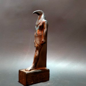 May include: A dark bronze statue of the Egyptian god Horus, depicted as a man with a falcon head. The statue is standing on a base with hieroglyphics.