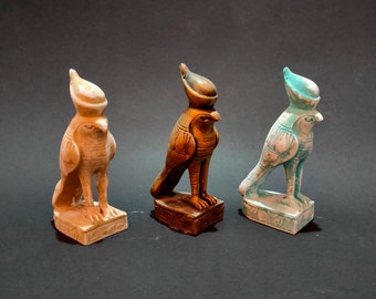 Puede incluir: Tres pequeñas figuritas decorativas del dios egipcio Horus, cada una con un esquema de color diferente: blanco, marrón y azul verdoso.