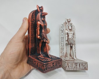May include: Two decorative statues of the Egyptian god Anubis, one in copper and one in white. Both statues are standing on a base with hieroglyphics.