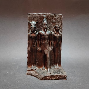 May include: A bronze-colored Egyptian relief sculpture depicting three figures. The central figure, a pharaoh, is flanked by two other figures, all in traditional attire. The sculpture has hieroglyphs at the base and is set against a textured background.