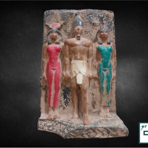 May include: A terracotta relief sculpture depicting three figures. The central figure is a male with a white headdress and a white kilt. The figures on either side are female, one wearing a red dress and the other wearing a teal dress.
