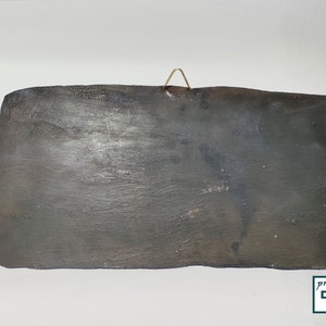 May include: A dark gray rectangular ceramic tile with a rough texture. The tile has a small metal hook on the top for hanging.