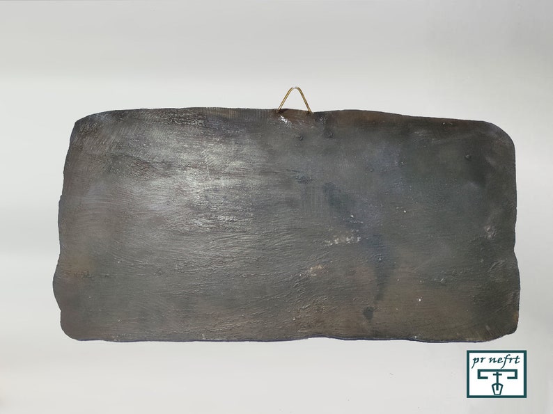 May include: A dark gray rectangular ceramic tile with a rough texture. The tile has a small metal hook on the top for hanging.