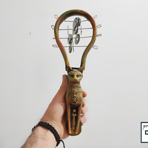 Op de afbeelding: Een handgehouden muziekinstrument in de vorm van een kat met een gouden afwerking en een set bekkens die aan een draadframe zijn bevestigd. De bekkens zijn van metaal en zijn in een cirkelvormige opstelling geplaatst.