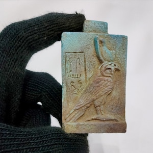 Puede incluir: Una pequeña placa rectangular de cerámica azul y marrón con un relieve tallado de un halcón con un disco solar en la cabeza. Jeroglíficos están tallados en el lado izquierdo de la placa.