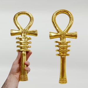 May include: Two gold Ankh symbols, an ancient Egyptian symbol of life, with a decorative, stacked, circular design on the shaft.