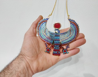 Puede incluir: Collar de tono dorado con un colgante de esmalte azul, rojo y turquesa que representa a la diosa egipcia Isis, con las alas extendidas. El colgante presenta un disco solar de esmalte rojo en la parte superior de la cabeza de la diosa.