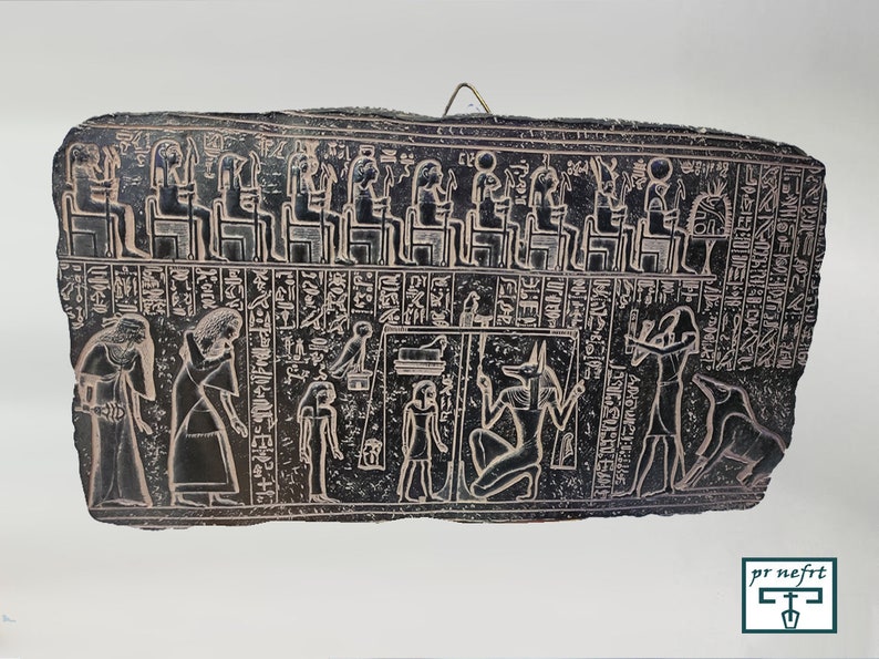 May include: Black stone relief carving depicting a procession of figures in front of a seated group. The figures are wearing traditional Egyptian clothing and headdresses. The seated group includes a figure with a jackal head, likely representing Anubis, the god of the dead.