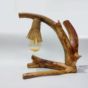 Puede incluir: Una lámpara de mesa de madera rústica con un diseño de rama natural. La lámpara tiene una pantalla de yute y una bombilla de vidrio transparente.