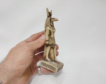 May include: A small, white, detailed figurine of the Egyptian goddess Bastet, with a cat head and human body, standing on a base with hieroglyphics.