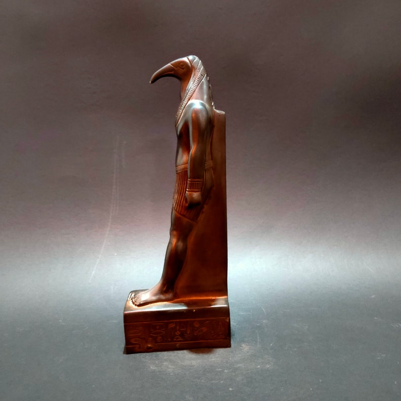 May include: A dark brown statue of the Egyptian god Thoth, depicted as a man with the head of an ibis. The statue is standing on a base with hieroglyphics.