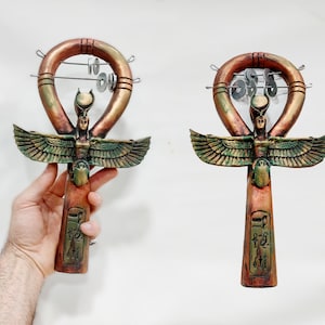 May include: Two Ankh-shaped decorative items with a bronze finish, featuring an Egyptian goddess figure with outstretched wings. The ankhs have a loop at the top with metal discs and hieroglyphic inscriptions on the base.