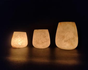 May include: Three glowing, off-white votive candle holders of varying sizes are arranged in a row. The holders have a rounded, organic shape and a textured surface. The warm light inside creates a soft, inviting ambiance.