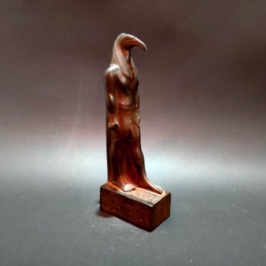 May include: A dark brown wooden statue of the Egyptian god Thoth, depicted as a man with the head of an ibis. The statue is standing on a rectangular base with hieroglyphics.