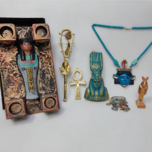 Può includere: Un set di oggetti decorativi a tema egizio che include una scatola di legno con una statuetta all'interno, un ankh dorato, uno scettro dorato con un uccello in cima, una statuetta blu e dorata, una collana blu e dorata con un ciondolo a forma di viso e una statuetta dorata.