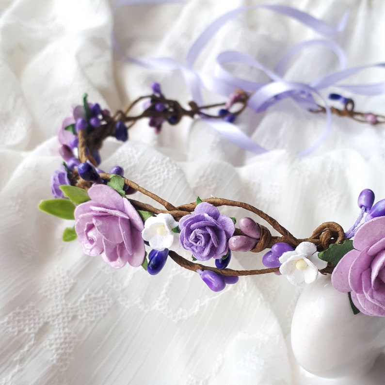 May include: A purple and white flower crown with a brown vine base. The crown is decorated with small purple flowers, white flowers, and purple berries.