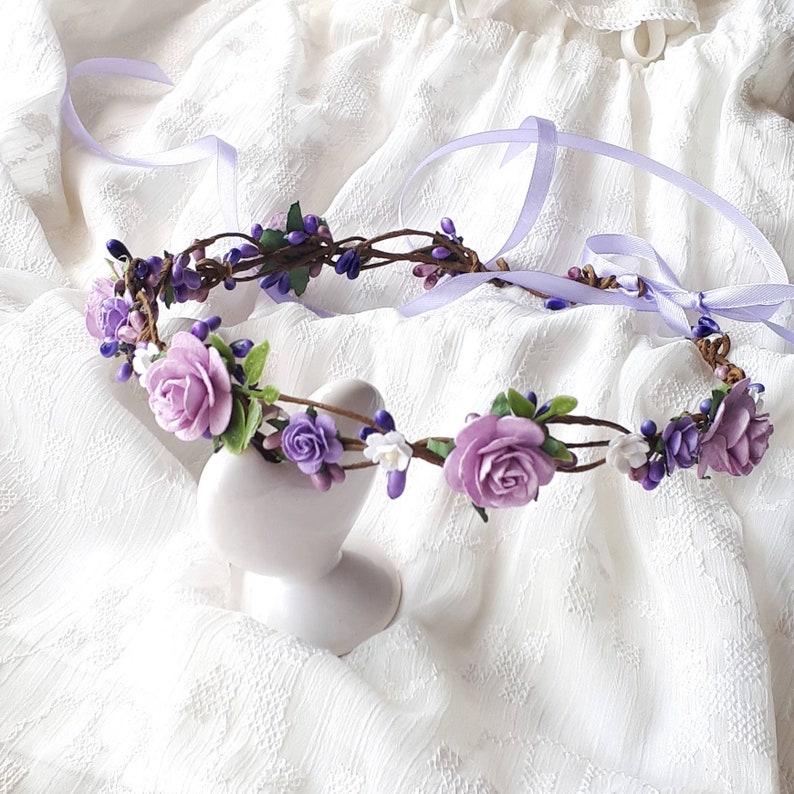 May include: A purple and white flower crown with a satin ribbon. The crown is made of artificial flowers and is displayed on a white heart-shaped stand.