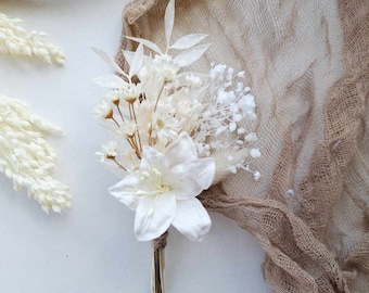 May include: A white dried flower bouquet with a single large white lily, small white flowers, and dried grasses. The bouquet is tied with twine and is sitting on a white surface with a brown burlap fabric in the background.