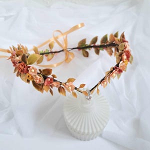 Puede incluir: Una corona floral con pequeñas flores melocotón y amarillas, hojas marrones y verdes, y una cinta dorada. Tiene un pequeño acento de cristal. La corona se exhibe sobre una superficie blanca texturizada. Ideal para una ocasión especial.