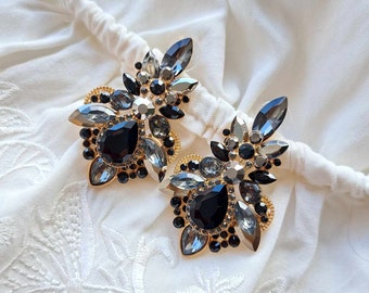 May include: A pair of large statement earrings with a cluster of black and clear rhinestones. The earrings have a gold tone finish and are designed in a floral shape.