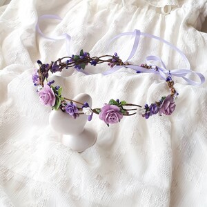May include: A purple flower crown with white flowers and green leaves. The crown is made of a brown vine base and is tied with a lavender ribbon.