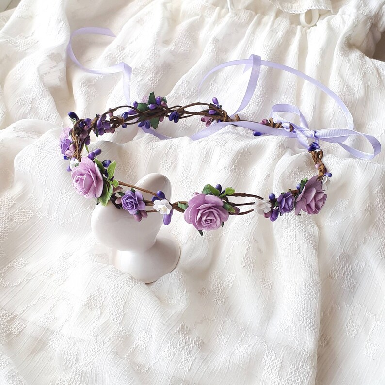 May include: A purple flower crown with white flowers and green leaves. The crown is made of a brown vine base and is tied with a lavender ribbon.
