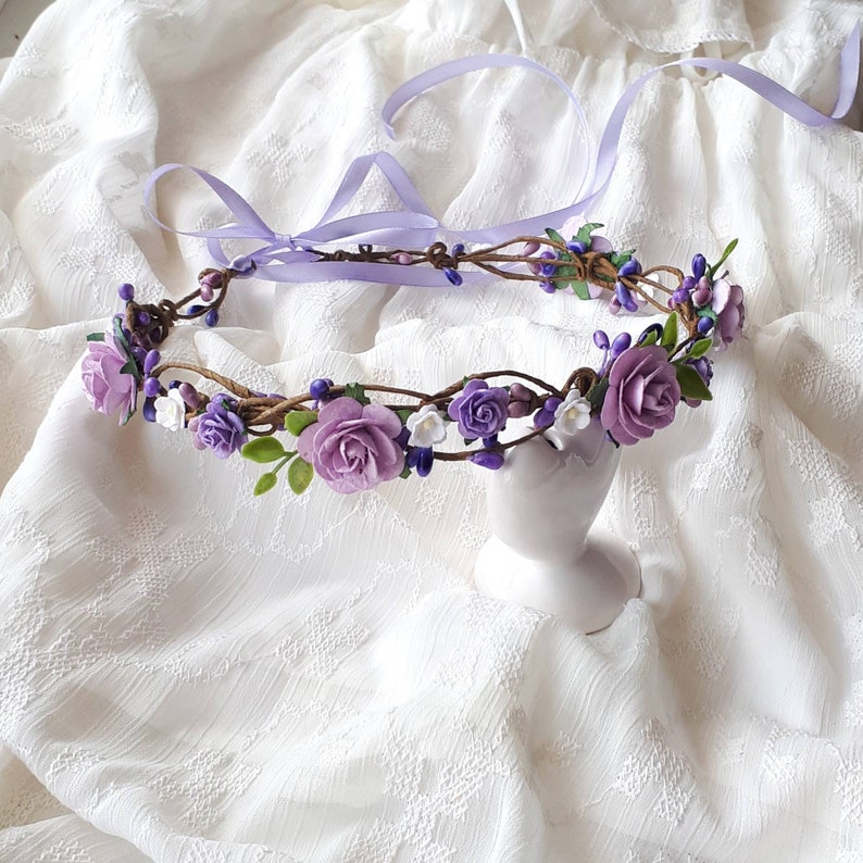 May include: A purple and white flower crown made of faux flowers and vines. The crown is sitting on a white fabric background.