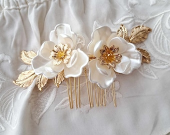 May include: Two white flower hair combs with gold-colored accents. Each comb features a large, pearlescent flower with a gold center and gold leaves. The combs have gold-colored teeth and are set against a white, textured background.