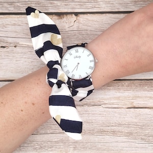Peut inclure: Une montre-bracelet avec un cadran blanc et un boîtier argenté. La montre a des chiffres romains et des aiguilles noirs. Le bracelet est en tissu rayé bleu marine et blanc avec des cœurs dorés. La montre est sur un poignet.