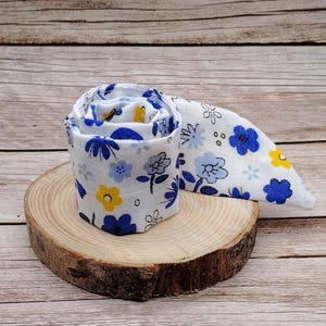 Può includere: Tessuto bianco arrotolato con un motivo floreale blu e giallo. Il tessuto è arrotolato e posto su una piccola fetta di legno. Il motivo floreale presenta fiori blu e gialli con steli e foglie verdi.