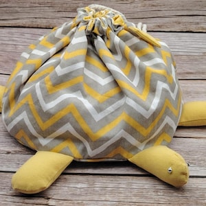 Pode incluir: Uma bolsa com cordão em forma de tartaruga com um padrão de chevron cinza, branco e amarelo. A cabeça e as quatro patas da tartaruga são amarelas. A bolsa é feita de tecido e está sobre uma superfície de madeira.
