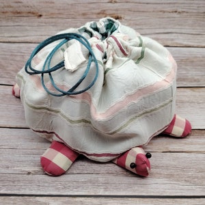 Peut inclure: Un sac à cordon en forme de tortue, en tissu blanc à rayures roses et vertes. Les pieds de la tortue sont en tissu à carreaux rouges et blancs. Le sac a un cordon de serrage turquoise et des yeux en perles noires.