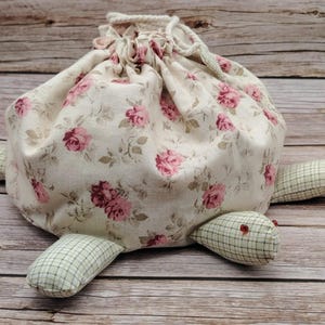 May include: A whimsical turtle-shaped storage bag. The body is a cream-colored fabric with pink rose floral patterns. The turtle's head and feet are a green and white checkered pattern. The bag has a drawstring closure.
