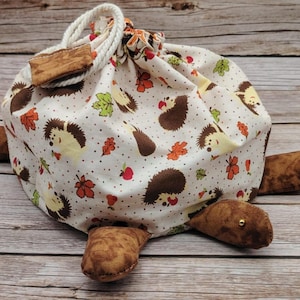 May include: A whimsical drawstring bag shaped like a turtle, featuring a white fabric with a repeating pattern of hedgehogs, leaves, and berries. The turtle's shell is the bag, and the feet are brown fabric. A wooden toggle and rope closure complete the design.