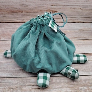 Peut inclure: Un sac à cordon de couleur vert sarcelle en forme de tortue avec des accents en tissu à carreaux blancs et verts. Le sac a une fermeture à cordon et une poignée.