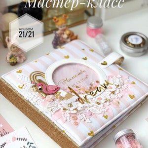 May include: A pink and white striped scrapbook with gold accents and a clear window with the text "Miroslava 01.12.2018". The scrapbook is decorated with pink and white flowers, a pink flamingo, and gold glitter.