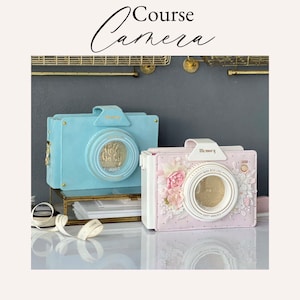 Può includere: Due borse a forma di macchina fotografica, una blu chiaro e una rosa con dettagli bianchi. Entrambe le borse hanno un'iscrizione dorata che recita "Memory". La borsa rosa ha un design floreale e il testo "Made with Love" e "Happy Moments".