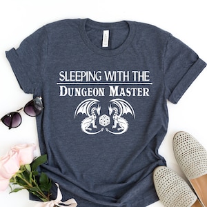Puede incluir: Una camiseta azul marino con texto blanco que dice "Sleeping with the Dungeon Master" y un gráfico de dos dragones enfrentados con un dado en el medio.