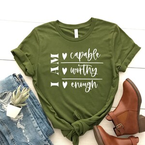 May include: A green t-shirt with the text "I AM" and three lines of text below it: "capable", "worthy", and "enough". Each line of text has a small heart symbol before it.