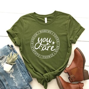 Könnte beinhalten: Ein grünes T-Shirt mit einem weißen Kreis-Motiv, auf dem "You are" steht, umgeben von den Worten "capable, enough, worthy, loved, kind, strong".
